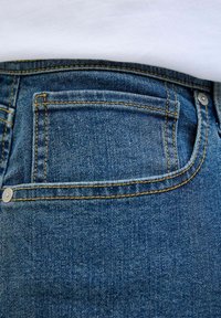 Primer plano de un bolsillo de vaqueros de mezclilla azul con costuras amarillas y remaches metálicos, sobre un fondo de tela blanca.