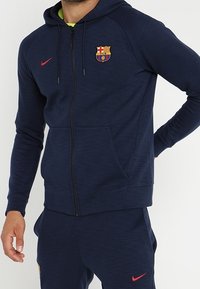 Felpa navy con zip realizzata in tessuto testurizzato, dotata di tasca frontale e di un patch con logo. Include un cappuccio e polsini a costine.