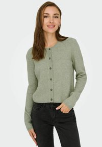 Gaiši zaļš kardigans, izgatavots no mīksta auduma, ar apaļu kakla izgriezumu, garām piedurknēm un pogu aizdari priekšpusē. Saskaņots ar melnām džinsām.