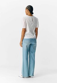 Witte blouse met pofmouwen en bloemenborduursel, met een sleutelgat aan de achterkant, gecombineerd met lichtblauwe wijde jeans, staand op een neutrale achtergrond.