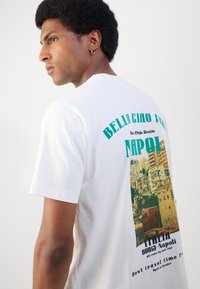 Witte katoenen T-shirt met een kleurrijke geprinte afbeelding van een stadsgezicht op de achterkant, met groene tekst die leest: “BELLA CIAO VAN NAPOLI.”