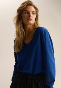 Niebieski sweter w serek z długimi rękawami, gładka faktura, w połączeniu z czarnymi spodniami w paski. Modelka ma długie, faliste włosy. Neutralne tło.