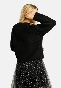 Melns adīts džemperis ar teksturētu ribveida dizainu, ar garām piedurknēm un īsu griezumu, kopā ar caurspīdīgu melnu svārku ar sirdīs rakstiem.