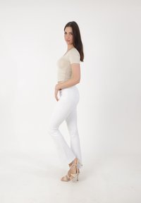 Woman with long brown hair wearing a beige fitted top, white flared pants, and beige high-heeled sandals, standing sideways against a white background.