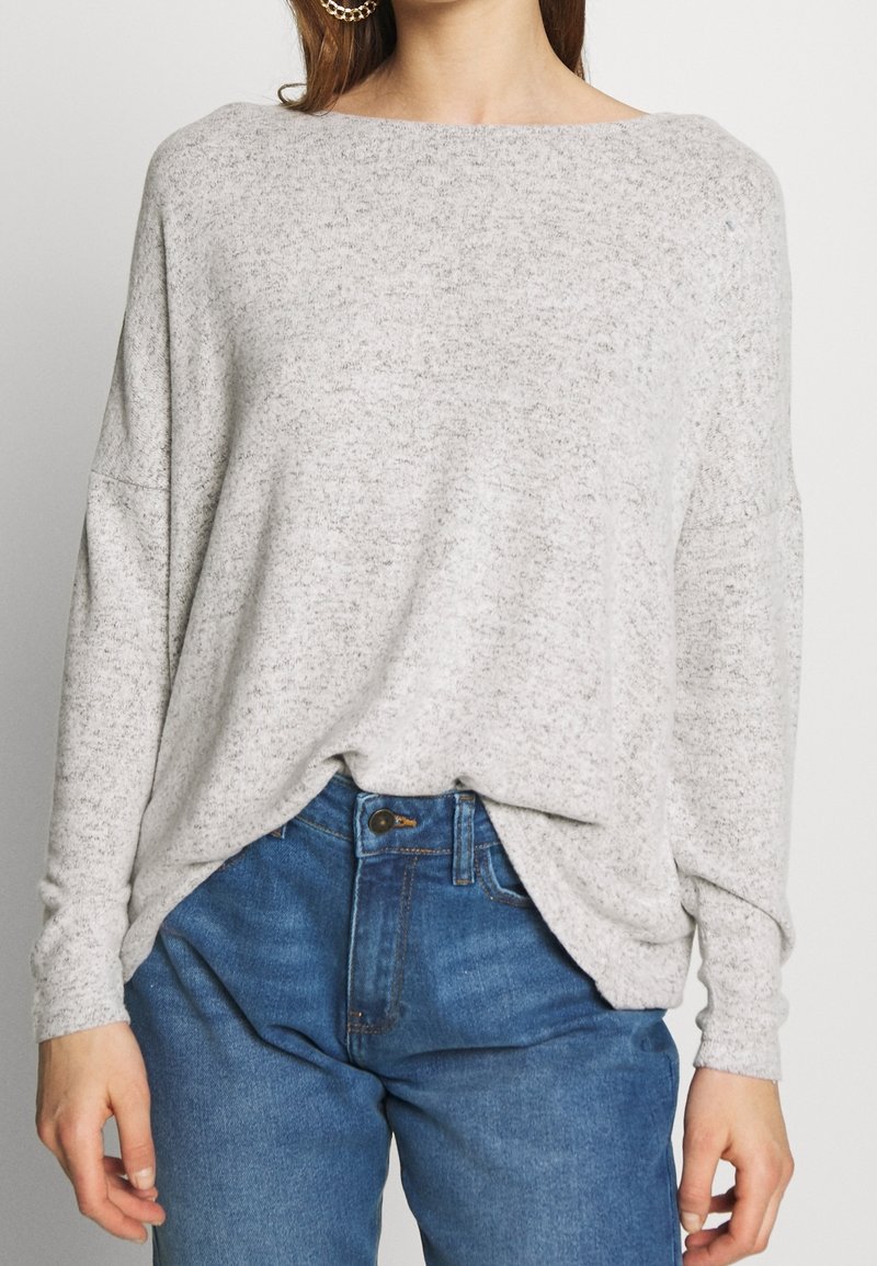 Světlý šedý pletený svetr s výstřihem do člunu, volným střihem a spadlými rameny, v kombinaci s modrými džínami z denimu. Měkká textura, minimalistický design.