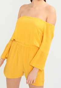 Gul off-shoulder jumpsuit med utsvängda ärmar, åtsittande midja och mjuk textur. Har en avslappnad passform och knälånga shorts.