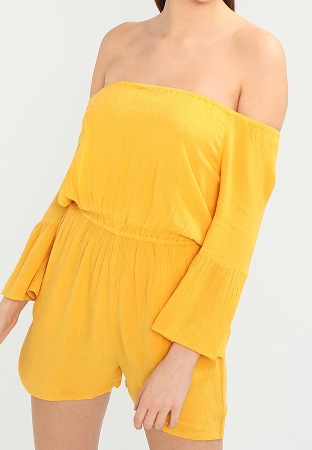 Gul off-shoulder jumpsuit med utsvängda ärmar, åtsittande midja och mjuk textur. Har en avslappnad passform och knälånga shorts.