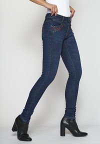 Kapeat farkut tummansinisestä denimistä, joissa on kukkakirjontaa takataskuissa. Farkuissa on korkea vyötärö ja ommeltuja yksityiskohtia.