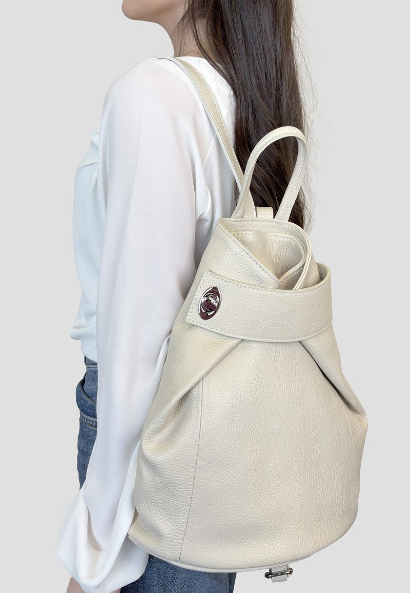 Mujer con blusa blanca y vaqueros azules lleva una mochila de cuero crema con solapa y broche plateado en el hombro.