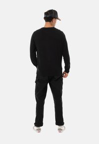 Chemise noire à manches longues, pantalons cargo noirs amples et baskets à carreaux. La personne porte une casquette noire avec un motif texturé.