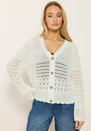 Lyshåret kvinde iført en hvid strikket cardigan med knapper og hulmønstre samt blå jeans, stående mod en ensfarvet lys baggrund.