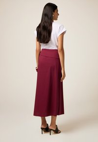 Falda maxi color burdeos con una cinturilla ajustada, hecha de una tela suave, combinada con una blusa blanca de manga corta y sandalias negras de tacón.
