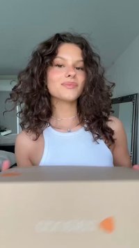 Gaišas kartona kaste ar izteiktu oranžu logotipu tiek turēta priekšā personai, kas valkā baltu bezpiedurkņu krekls un delikātu kaklarotu.