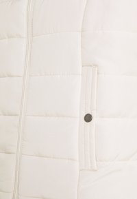 Jasnobeżowa kamizelka puchowa z poziomym pikowaniem, zamek błyskawiczny z przodu, wzmocnione szwy oraz detale z zatrzaskowymi kieszeniami.