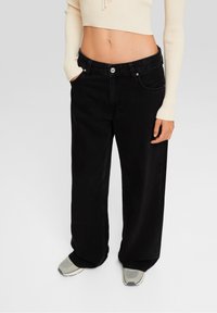 Jeans de pierna ancha negros fabricados en denim, con bolsillos frontales, cierre de botón y un corte relajado. Combinados con una camiseta corta de canalé en color crema.