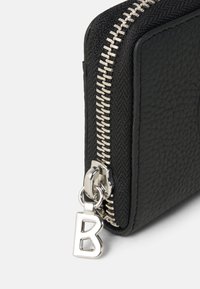Monedero negro hecho de cuero texturizado con una cremallera plateada. Presenta un encanto plateado con la letra "B" enganchado al tirador de la cremallera.