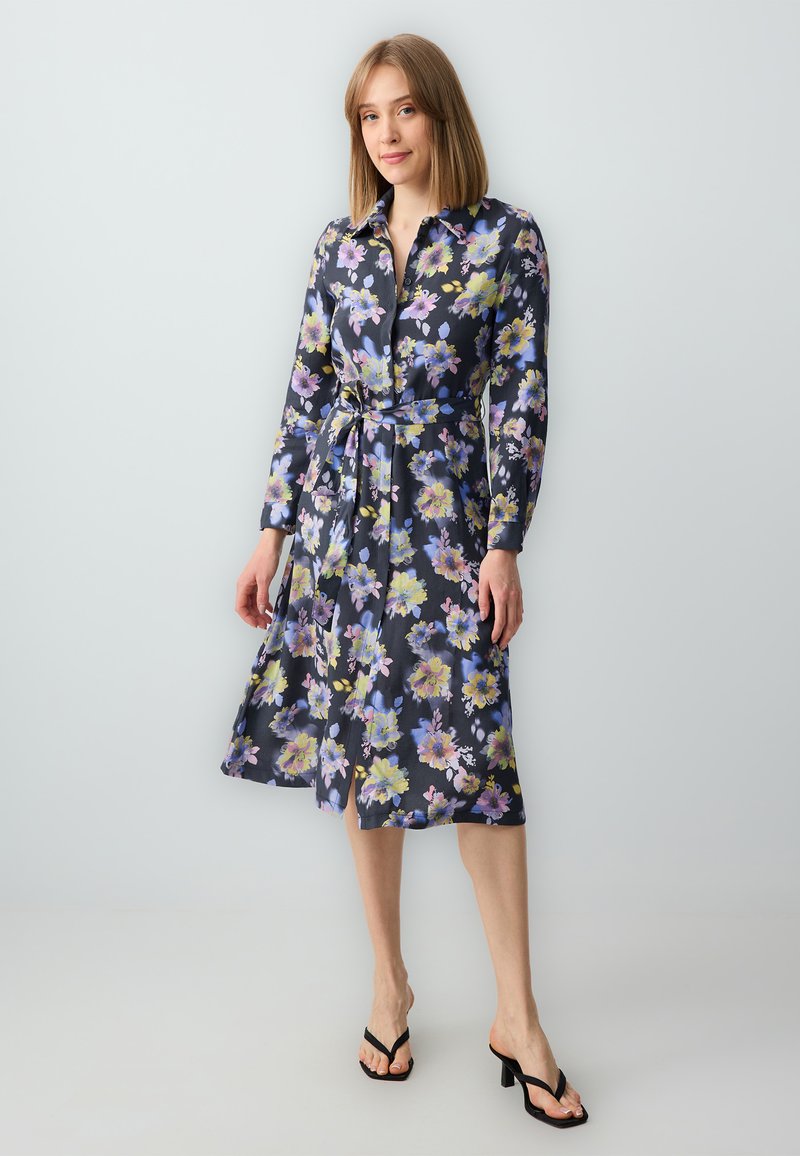 Blumenhemdkleid mit langen Ärmeln, dunkelblauem Grund, lebhaften gelben und lila Blumen, tailliert, knielang, begleitet von schwarzen Sandalen.