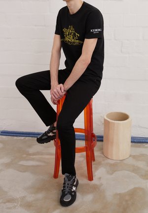 Mann trägt ein schwarzes T-Shirt mit Grafik und schwarze Hose, sitzt auf einem transparenten orangenen Barhocker auf Betonboden.