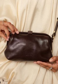 Mani che tengono una borsa clutch Balagai in pelle marrone scuro su uno sfondo di tessuto crema, con una mano che indossa due anelli distintivi.