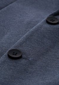 Tamno plava tkanina s suptilnom teksturom, s crnim dugmetom s četiri rupe i uzdignutim natpisom. Pogled izbliza na detalj odjeće.