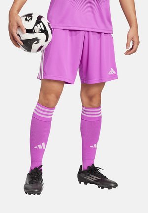 Roze voetbalkorte broeken en sokken hebben witte strepen en een logo. Zwarte voetbalschoenen met een gestructureerde oppervlakte maken de sportoutfit compleet. De speler houdt een voetbal vast.