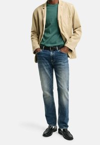 Beige Steppjacke über einem grünen T-Shirt, kombiniert mit blauen, ausgebleichten Jeans und schwarzen Slippern. Auffällige Tasche an der Jacke.
