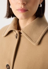 Manteau en laine beige avec un grand col, doté de deux boutons. Sa texture douce et son design ajusté mettent en avant une simplicité moderne.