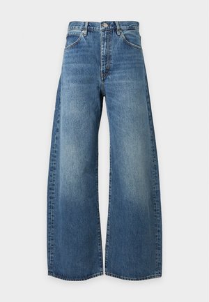 Femme portant un polo beige à manches longues, jean bleu à jambes larges, talons pointus noirs, tenant un sac à main noir sur un fond uni.