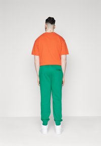 Orange bomullströja och gröna sweatpants med elastiska band, bärs med vita sneakers. Baksidan av outfiten syns mot en enfärgad bakgrund.