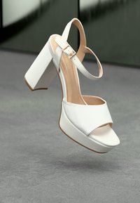 Sandalias de plataforma blancas con un tacón grueso, punta abierta y correa en el tobillo con un hebilla decorativa. Material suave con un acabado mate.