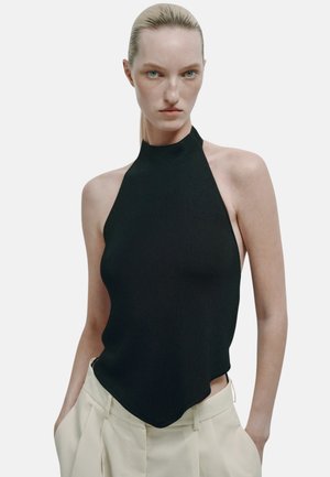 Blonde Frau, die ein ärmelloses schwarzes Neckholder-Top und hellbeige Hosen trägt, steht vor einem schlichten weißen Hintergrund.