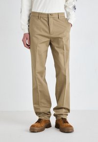 Beige pants with a straight leg design, featuring a zip-fly closure and button. Worn with brown lace-up shoes. Smooth texture, no patterns.