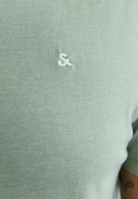 Világoszöld, pamutból készült póló, bal oldalon egy kis fehér hímzett „és” jellel. Simább textúrájú és kerek nyakú.