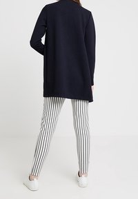 Marinblå lång cardigan med diagonalt ribbat mönster, matchad med svart och vit randiga leggings och vita sneakers, sedd bakifrån.