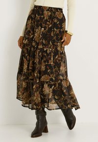Jupe maxi noire avec un motif floral en vert, rouge et or. Présente un design volé et est assortie à des bottes marron montantes.