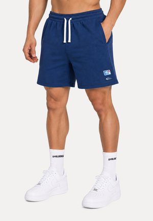 Man draagt marineblauwe shorts met trekkoord, witte sokken met merknaam en witte sportsneakers, staand tegen een effen achtergrond.