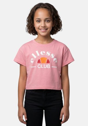 Jeune personne aux cheveux bouclés sourit, portant un t-shirt court rose avec le logo « ellesse CLUB » et un pantalon noir sur un fond uni.