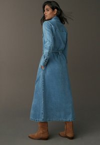 Robe en denim de couleur bleu clair, longue, cintrée à la taille avec un col. Elle dispose de poches et présente une coupe droite. Associée à des bottes en daim marron mi-mollet.