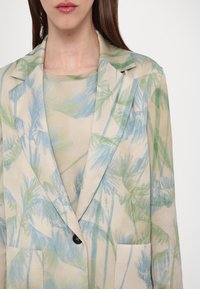 Femme portant un blazer beige et un haut avec des motifs abstraits de feuilles bleues claires et vertes, boutonné à l'avant, visible du cou à la taille.