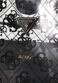 Matériau à motifs noir et blanc avec un design de logo répété, comprenant une étiquette métallique triangulaire avec "GUESS U.S.A." et un point d'interrogation.