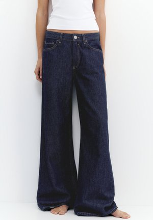 Person iført mørkeblå bukser med vide ben og en hvid ærmeløs top, stående barfodet mod en hvid baggrund.