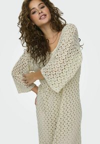 Vestido de encaje en color crema, con mangas anchas y sueltas y un patrón de ganchillo. El diseño es ligero y con un corte relajado.