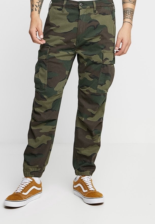 Personne portant un pantalon cargo camouflage vert et marron avec des poignets élastiques et des baskets marron avec des lacets blancs.