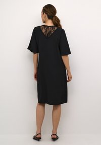 Schwarzes Kleid mit kurzen Ärmeln, das über einen Spitzenrücken-Einsatz und eine vertikale Nadelstreifenstruktur verfügt. Knielanger Schnitt mit lockerer Passform.