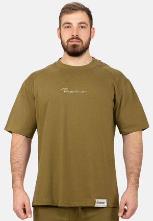 Bärtiger Mann trägt ein olivgrünes Oversize-T-Shirt mit kleinem weißem Logo auf der Brust und einem Etikett nahe dem Saum.