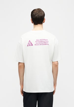 Persona vistiendo una camiseta blanca de manga corta con el logo y texto de Nike ACG en púrpura en la parte superior de la espalda, de pie contra un fondo claro y liso.