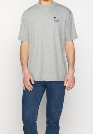 Homme portant un t-shirt Lacoste gris clair avec un petit logo sur la poitrine gauche, associé à un jean bleu, debout devant un fond uni.