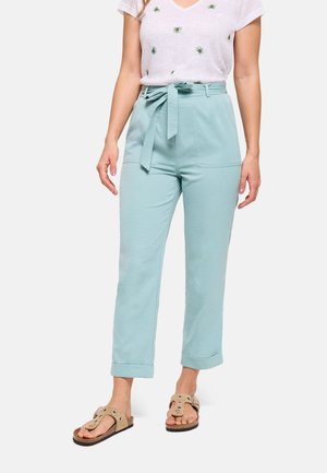 Mujer que lleva pantalones azul claro de cintura alta con cinturón atado, camiseta blanca con pequeños diseños verdes y sandalias beige abiertas en los dedos.