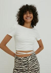 Weißes geripptes, kurzes T-Shirt mit kurzen Ärmeln, das ein Bindedetail um die Taille hat, kombiniert mit schwarz-weißen gestreiften Jeans.