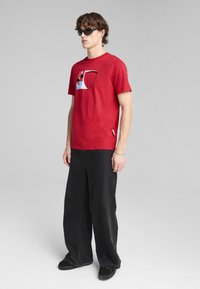 Camiseta roja de algodón con un diseño gráfico que presenta formas geométricas en varios colores. Combinada con pantalones de pierna ancha negros y zapatos oscuros.